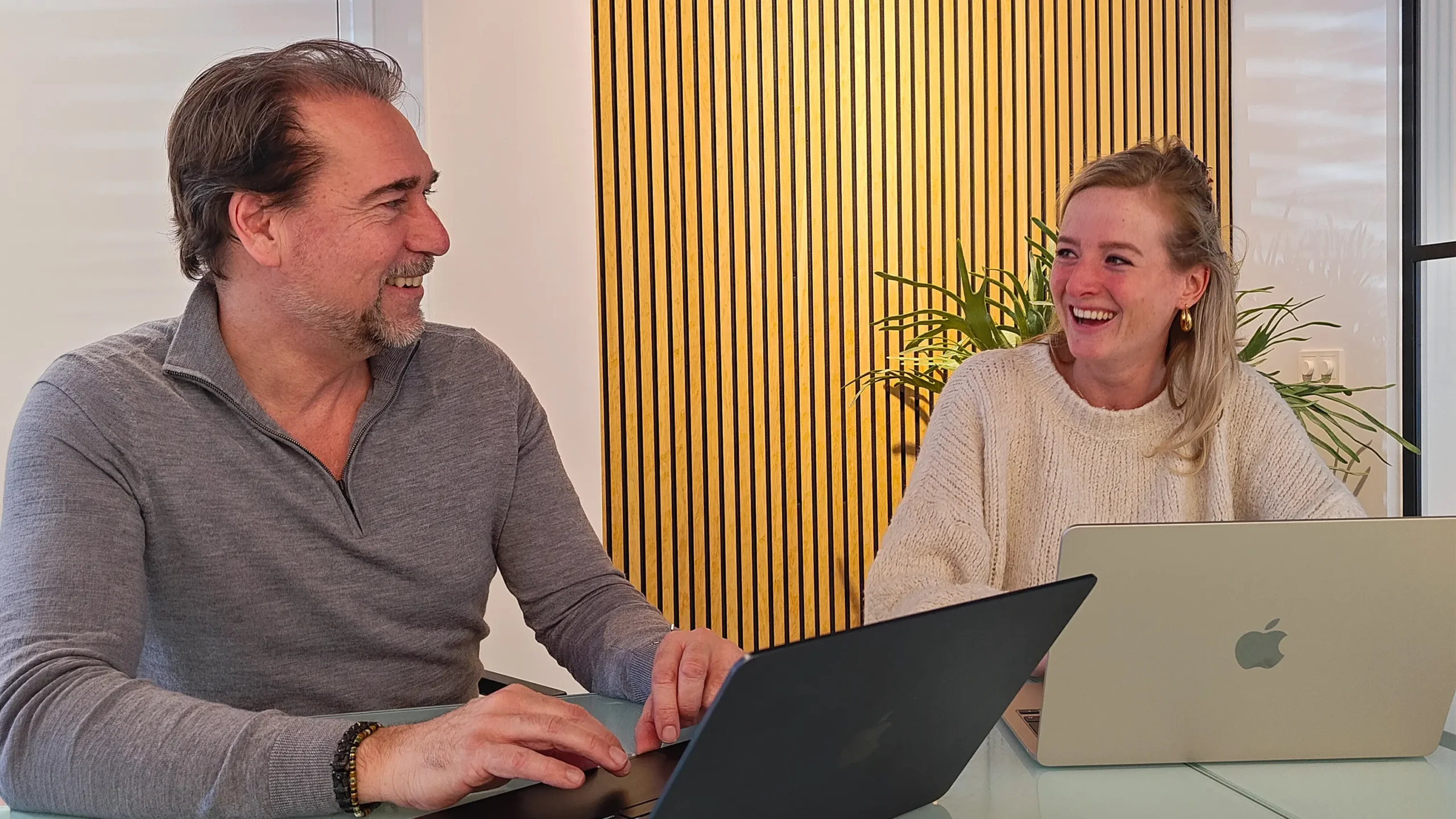 Twee collega’s lachen samen aan een tafel met laptops in een kantooromgeving