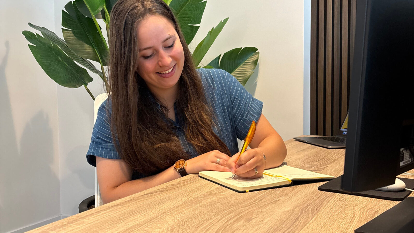 Meisje in blauwe blouse en lang donkerbruin haar achter bureau die in een schrift schrijft op kantoor over ux/ui design